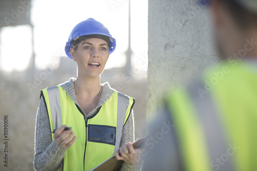 Tablou pe pânză Female surveyor with clipboard talking to builder on construction site