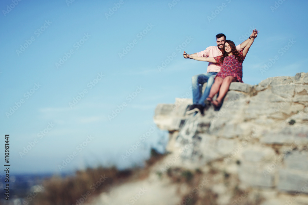 Happy free couple holding hands sitting on cliff, clear sky back