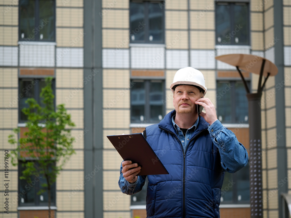 Engineer standing on the background of a new apartment building. House is built. Engineer in a helmet with papers in hand on the phone. The concept of new housing construction, real estate Engineer standing on the background of a new apartment building. House is built. Engineer in a helmet with papers in hand on the phone. The concept of new housing construction, real estate