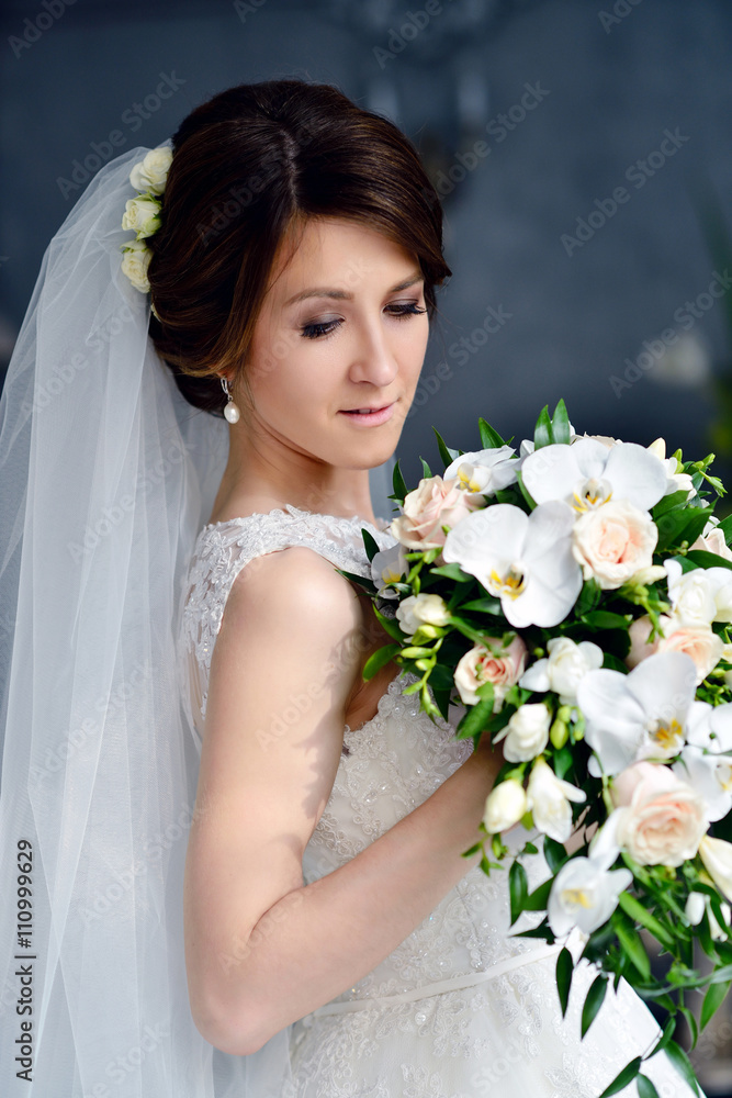 Naklejka premium Beauty bride in bridal gown with bouquet and lace veil indoors. Beautiful model girl in a white wedding dress. Female portrait of cute lady. Woman with hairstyle
