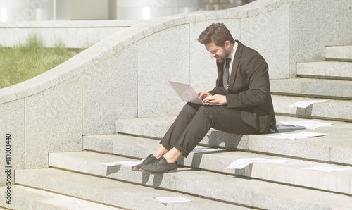 Toned picture of businessman in black business suit using laptop computer while sitting n city centre. Freelnace concept.