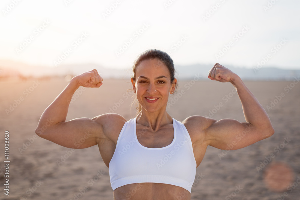Young fitness woman flexing big strong biceps muscles towards the sun at  urban beach. Cheerful female bodybuilder showing arms. Workout success  concept. Photos