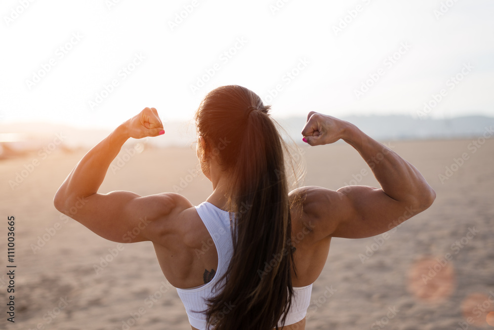 Fotografia do Stock: Young fitness woman flexing big strong biceps muscles  towards the sun at urban beach. Back view of female bodybuilder showing arms.  Workout success concept.