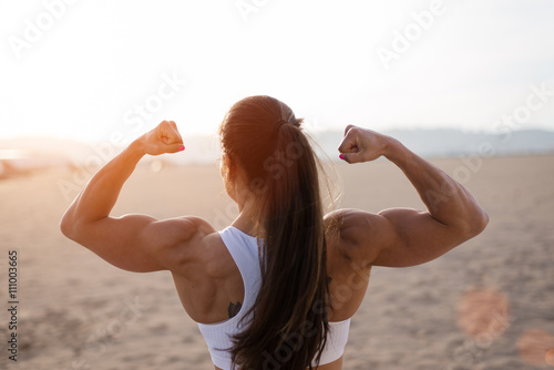 Obraz na plátně Young fitness woman flexing big strong biceps muscles towards the sun at urban beach