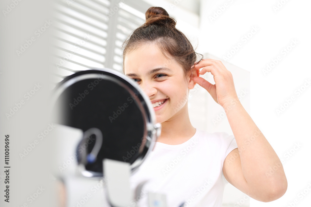 Niewidoczny aparat słuchowy. Wesoła dziewczynka zakłada aparat słuchowy przeglądając się w lusterku - obrazy, fototapety, plakaty 