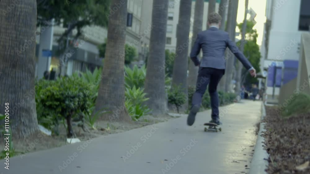 Young man in business suit rides away from the camera on a skateboard against a city background with traffic and trees in Downtown Los Angeles. Real time recorded at 24fps. Young man in business suit rides away from the camera on a skateboard against a city background with traffic and trees in Downtown Los Angeles. Real time recorded at 24fps.