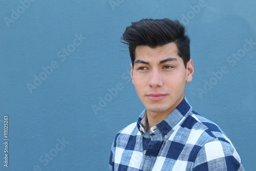 Handsome boy looking away leaning back against blue wall. Young hipster boy dressed in cool plaid shirt lost in thought. Street fashion look, a lot of space for copy 