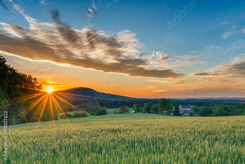 A perfect Spring day ends as the sun sets and sprays warm golden light over a beautiful farm with fields sprouting, a stone house, and mountain ridges in the background. photo