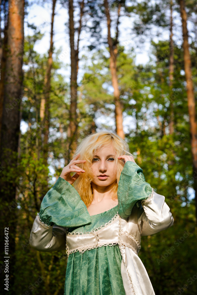 Fashionable shooting of a young short haired blond woman posing in the forest park wearing fancy empire style dress holding flowers. Concept of fantasy and magic. Fashionable shooting of a young short haired blond woman posing in the forest park wearing fancy empire style dress holding flowers. Concept of fantasy and magic.