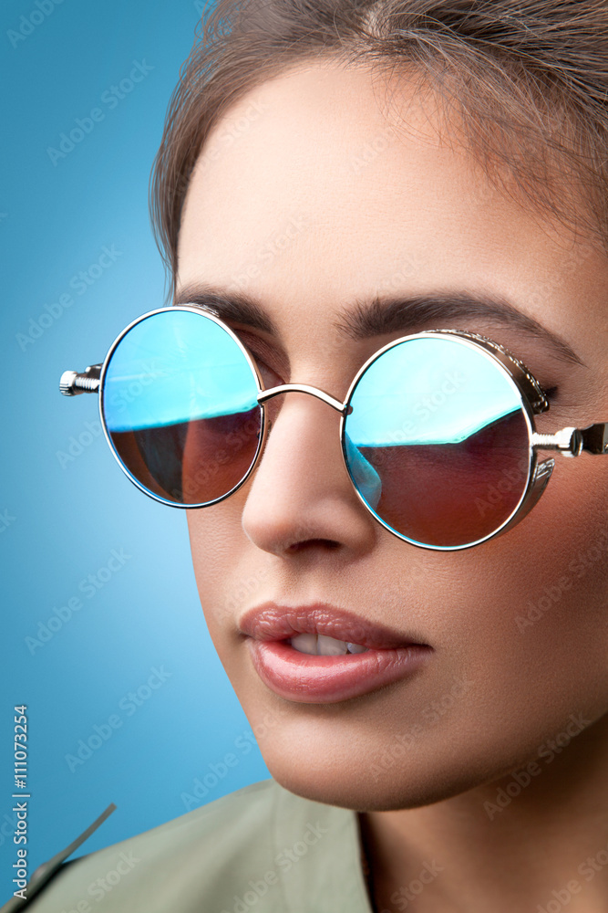 Close-up face portrait of young beautiful woman with perfect skin in round sunglasses smiling and looking at camera on the blue background. Beauty face make-up. Close-up face portrait of young beautiful woman with perfect skin in round sunglasses smiling and looking at camera on the blue background. Beauty face make-up.