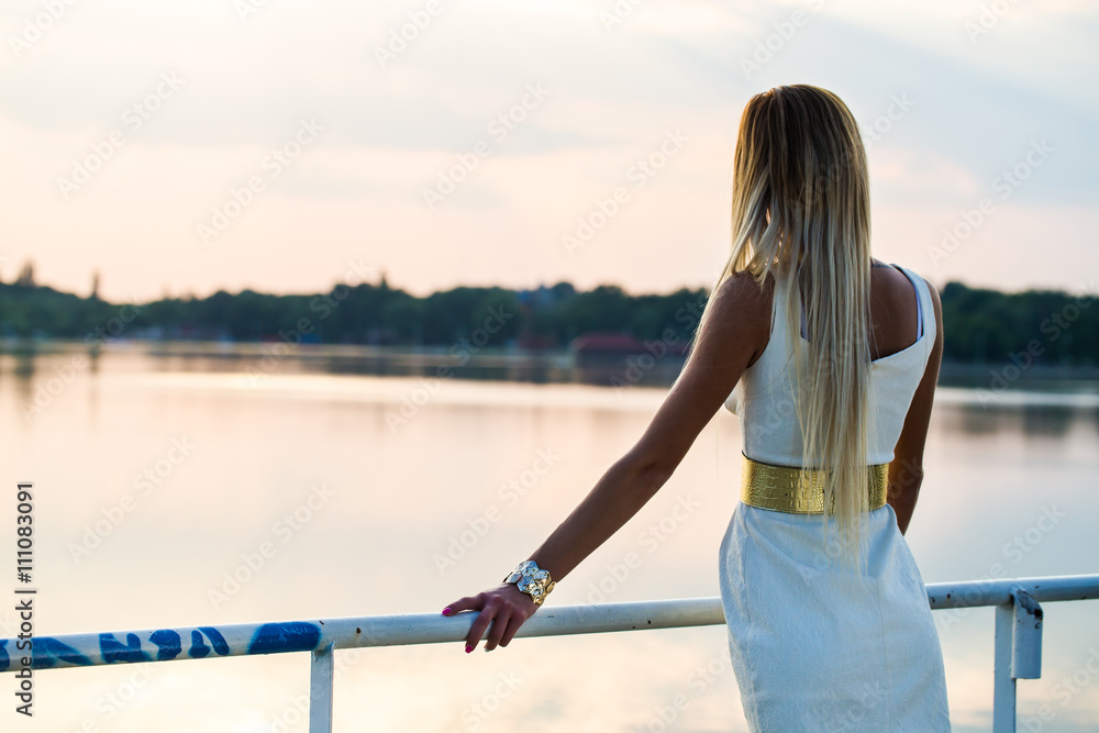 pretty dressed young girl looking to the lake from handrail at sunset