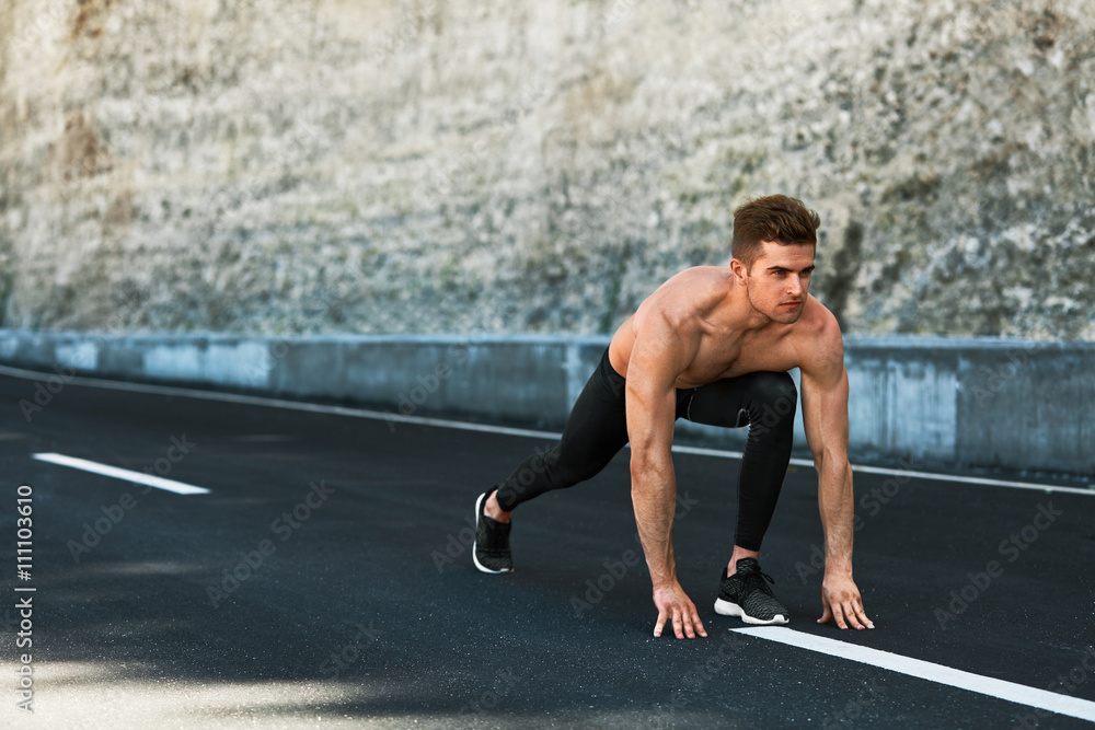 Fototapeta premium Sports. Healthy Athletic Man With Fit Muscular Body In Starting Position For Running On Road. Handsome Runner Ready To Start Sprint Race. Fitness Model Training Outdoors In Summer. Workout Concept