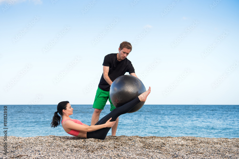 Sport und Fitness Instruktor zeigt junger Frau wie man mit Gymnastikball den Bauch trainiert am Strand im Freien - obrazy, fototapety, plakaty 