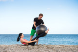 Sport und Fitness Instruktor zeigt junger Frau wie man mit Gymnastikball den Bauch trainiert am Strand im Freien
