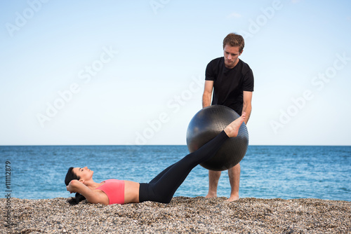 Sport und Fitness Lehrer zeigt junger Frau wie man mit Gymnastikball den Bauch trainiert am Strand im Freien