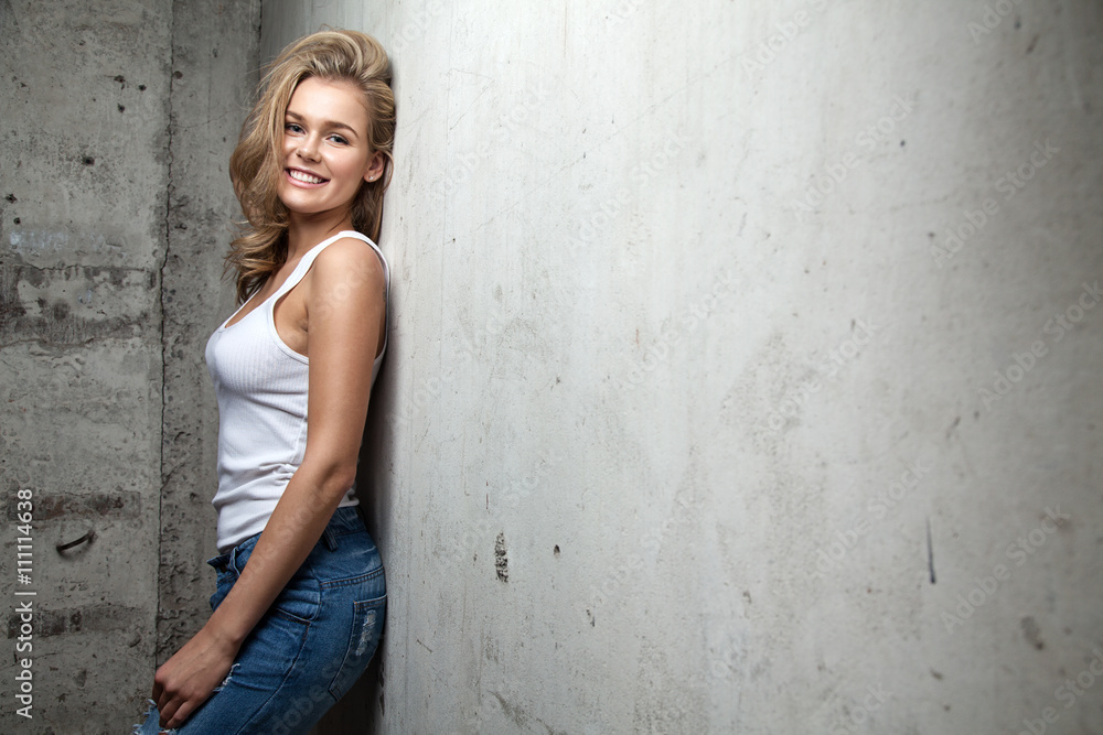 Cheerful blonde with natural beauty in a white shirt and blue jeans standing on the cement wall background