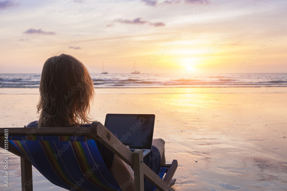 Young female traveler using laptop computer on the beach, sunset