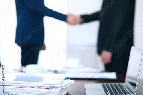 Documents and laptop on the table. Business people shaking hands on the background