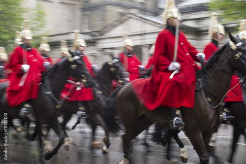 Royal guards on horseback dressed in ceremonial red coats pass in a parade on a rainy day in London, England, UK
