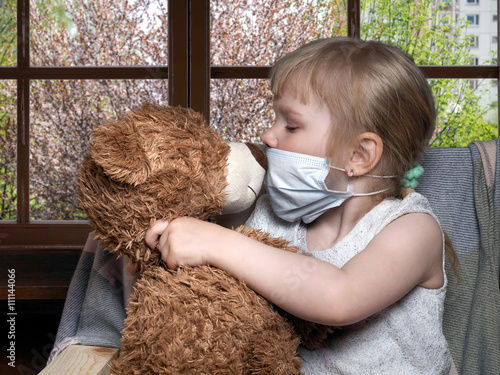 Little kid in a medical mask kissing toy bear. Outside, the trees are blooming. Conceptually about allergies, colds and Ecology