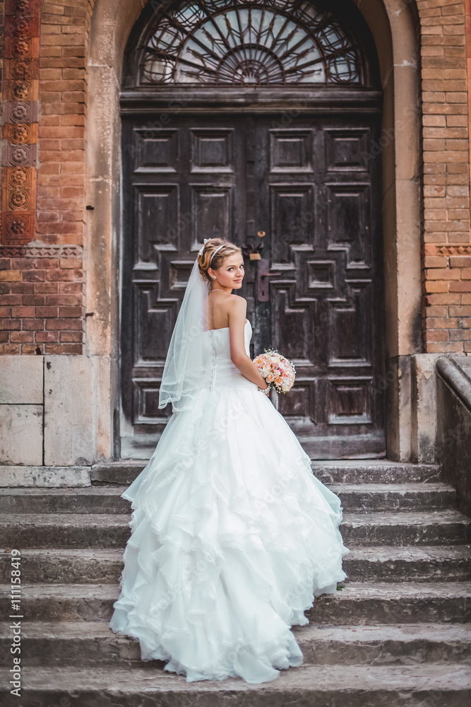 Fototapeta premium Beautiful blond bride in white dress posing near old wooden door