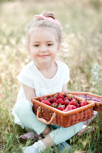 Cute baby girl 3-4 year old having picnic outdoors. Eating fresh strawberry. Looking at camera.