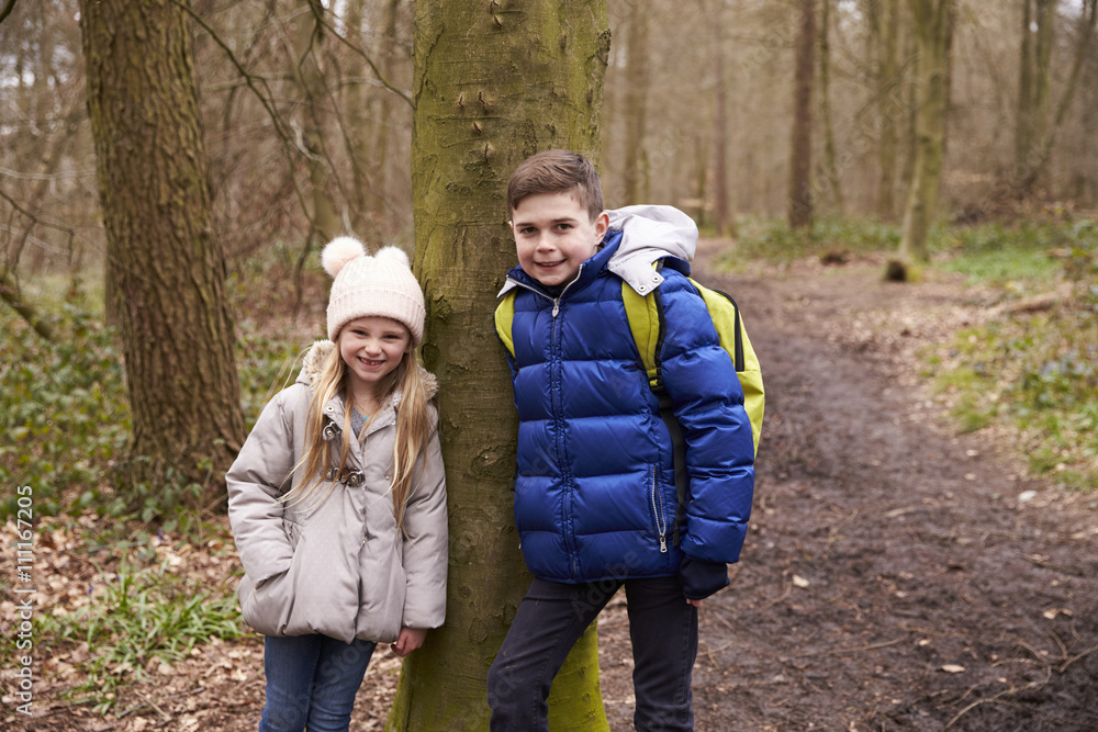 Brother and sister lean against a tree by a path in a forest