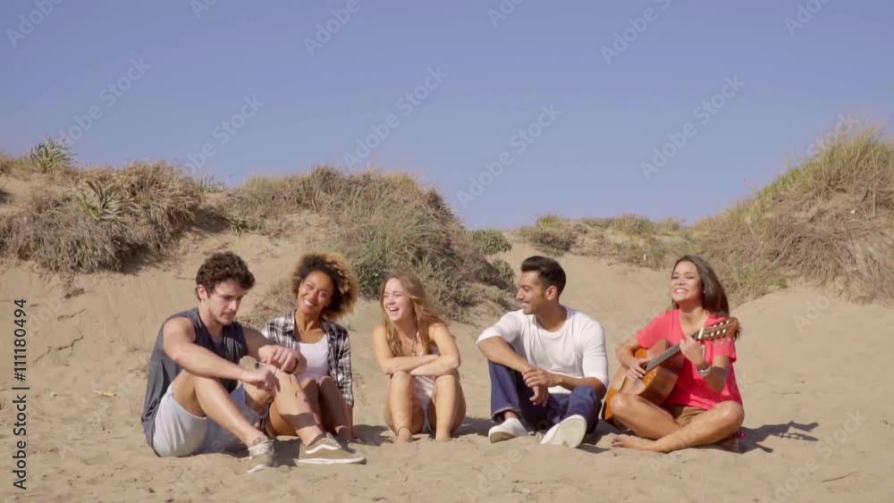 Five friends sitting on the sandy beach and listening acoustic guitar. Young brunette woman wearing in red blouse playing acoustic guitar for friends on the beach in slow motion. Five friends sitting on the sandy beach and listening acoustic guitar. Young brunette woman wearing in red blouse playing acoustic guitar for friends on the beach in slow motion.