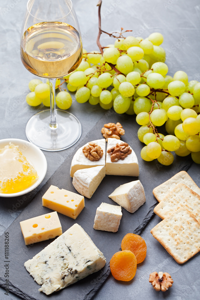 Glass of white wine and assorted cheese with grape on a stone and slate background.
