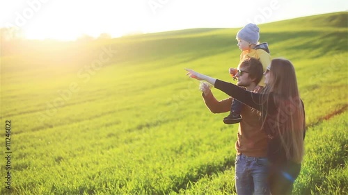 Happy joyful smiling young parents family father mother and little son on fathers shoulders walking on green grass meadow in countryside pointing at smth having fun outdoors in nature - slow motion  photo