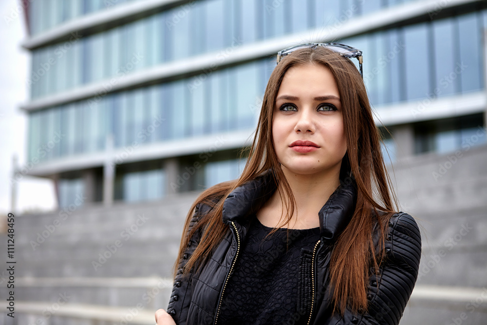 Fototapeta premium Fashion concept. Beautiful young woman in nice black coat. Posing on glass building. City lifestyle. Female fashion. Closeup portrait