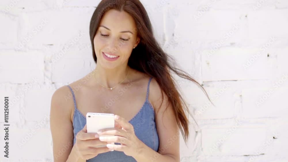 Pretty young woman leaning back against a white stone wall with her hair blowing in the breeze checking a text message on her smartphone Pretty young woman leaning back against a white stone wall with her hair blowing in the breeze checking a text message on her smartphone