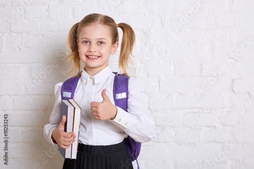Cute smiling schoolgirl in uniform standing with books and showing ok on light  background.