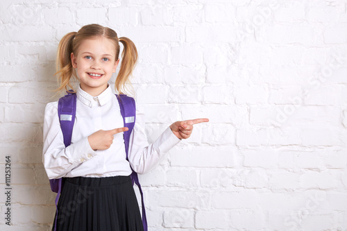 Cute smiling schoolgirl in uniform standing on light  background and showing  thumbs to the side. Copy space