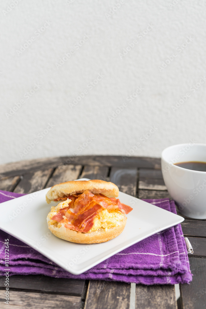 Salt muffin with scrambled eggs, bacon and cheese on white plate with dark coffee in white mug lying on wooden background. Unhealthy breakfast with bacon, eggs, pastry and coffee on purple dishtowel. Salt muffin with scrambled eggs, bacon and cheese on white plate with dark coffee in white mug lying on wooden background. Unhealthy breakfast with bacon, eggs, pastry and coffee on purple dishtowel.