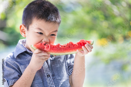 Cute little asian boy eating watermelon in park with sunshine