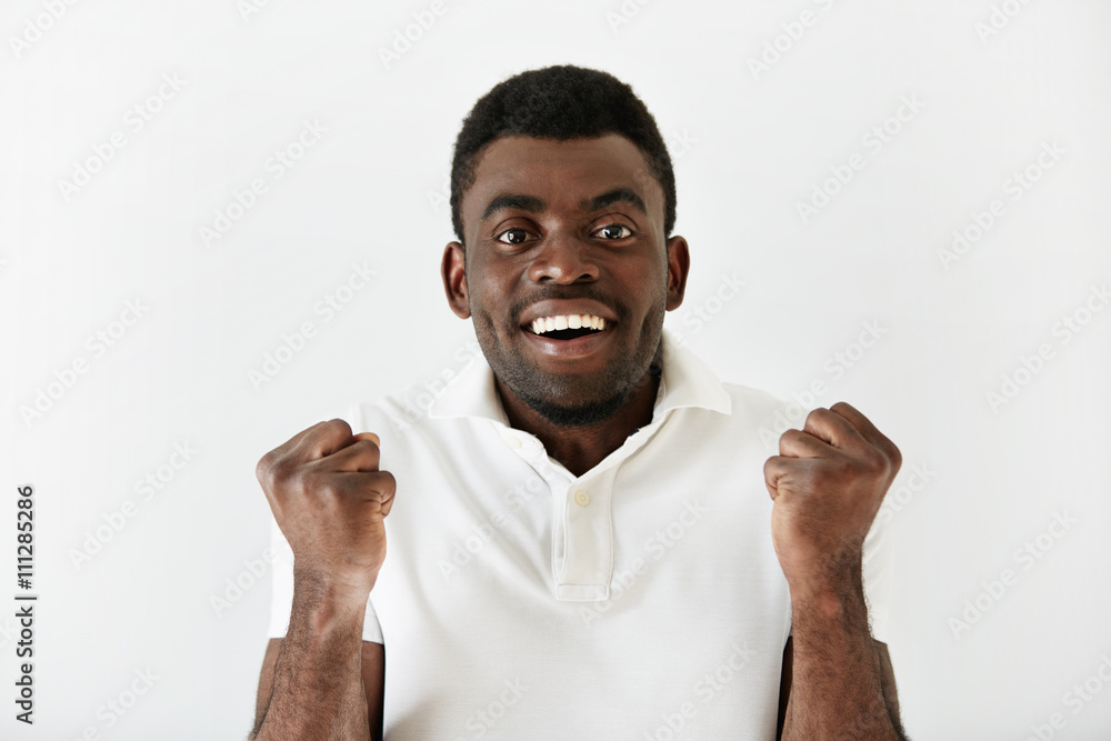 Headshot of happy successful African American student or businessman looking with winning expression, fists pumped, celebrating success against white wall background. Life perception, achievement Headshot of happy successful African American student or businessman looking with winning expression, fists pumped, celebrating success against white wall background. Life perception, achievement