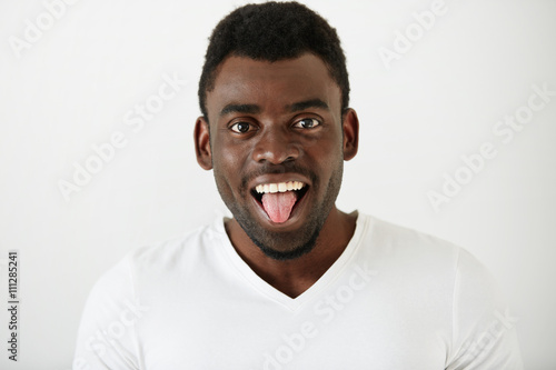 Close up portrait of African American student boy wearing white T-shirt making funny face, looking and showing tongue at the camera after classes at university. Attractive young male model having fun