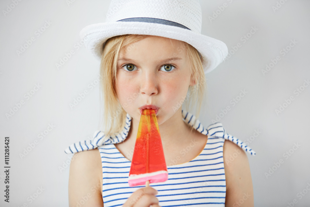 Isolated shot of cute little girl with green eyes and blonde hair, wearing white hat and striped dress, holding fruit ice cream. Portrait of Caucasian child licking popsicle and looking at the camera Isolated shot of cute little girl with green eyes and blonde hair, wearing white hat and striped dress, holding fruit ice cream. Portrait of Caucasian child licking popsicle and looking at the camera