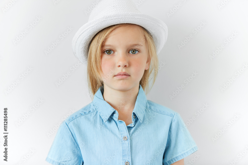 Close up studio portrait of beautiful Caucasian little girl with long blonde hair wearing white hat and denim shirt looking with serious expression at the camera. People and lifestyle concept. Close up studio portrait of beautiful Caucasian little girl with long blonde hair wearing white hat and denim shirt looking with serious expression at the camera. People and lifestyle concept.
