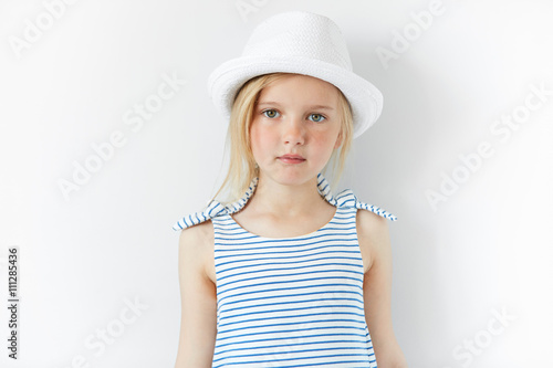 People and lifestyle concept. Close up of happy little female model posing against white concrete wall, wearing white hat and striped dress. Adorable blonde preschool girl looking at the camera