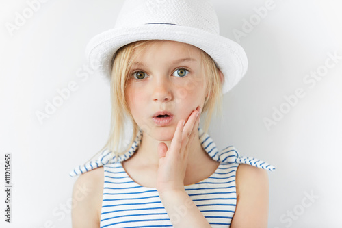 Portrait of surprised or frightened girl looking at the camera with a hand on her cheek. Close up shot of blonde Caucasian little girl with scared or shocked expression against white studio wall