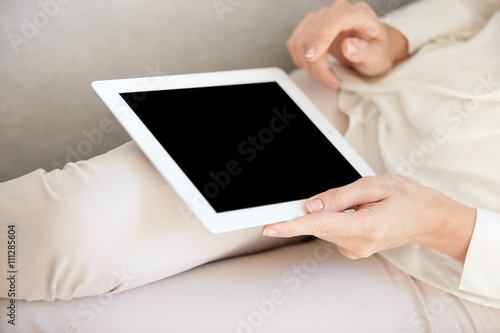 Cropped portrait of young woman using digital tablet at home, relaxing on comfortable sofa in the living room. Student girl messaging her friends and inviting them for the party. Selective focus
