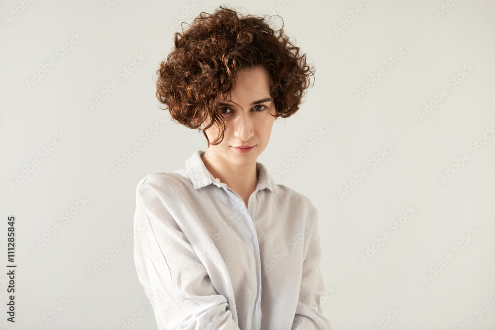 Close up of serious pretty Caucasian female with short brown curly hair wearing trendy white shirt. Fashion studio portrait isolated against white background with copy space for your advertisement. Close up of serious pretty Caucasian female with short brown curly hair wearing trendy white shirt. Fashion studio portrait isolated against white background with copy space for your advertisement.