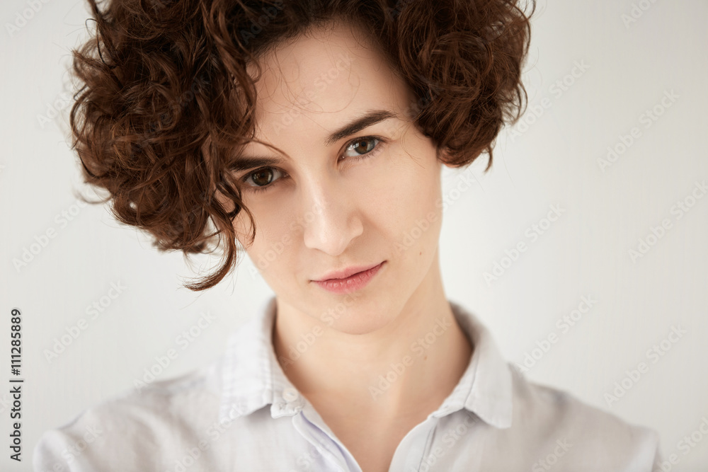 Close up shot of young brunette Caucasian woman looking with serious expression at the camera. Portrait of displeased or angry female office worker posing against white concrete wall background Close up shot of young brunette Caucasian woman looking with serious expression at the camera. Portrait of displeased or angry female office worker posing against white concrete wall background