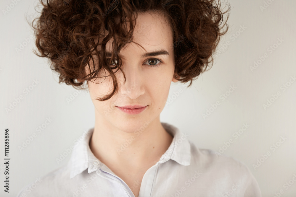 Close up shot of young beautiful Caucasian female looking and smiling at the camera. Hipster girl with natural makeup and curly hair wearing casual pastel clothes posing against white studio wall.