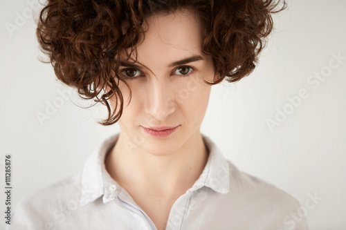 Close-up portrait of serious-looking Caucasian brunette woman with healthy and clean skin. Portrait of sensual young curly model with seductive eyes posing against white studio wall background