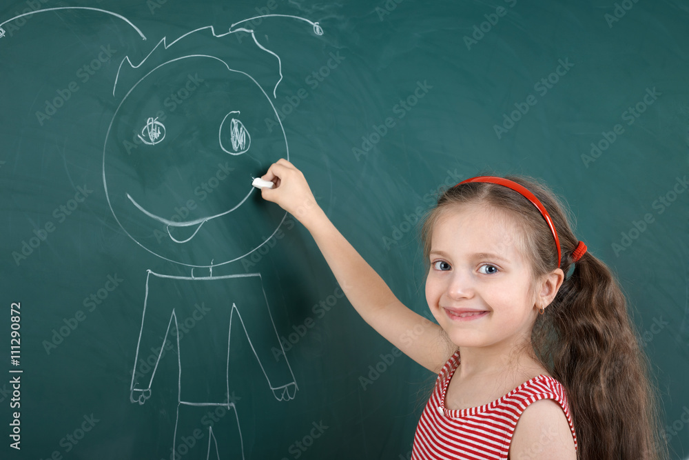 schoolgirl child in red striped dress drawing happy man on green chalkboard background, summer school vacation concept