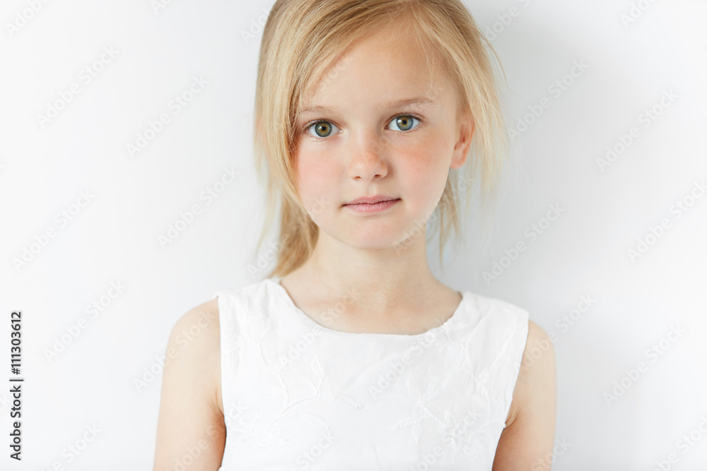 Close up shot of cute fashionable child with green eyes and blonde hair  weraing white dress, looking at the camera with serious expression.  Portrait of adorable little girl. Happy childhood concept Stock