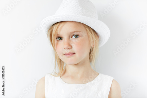 Lifestyle and people concept. Isolated headshot of beautiful Caucasian 5-year old female model with fair hair wearing stylish white clothes, looking and smiling at the camera with happy expression